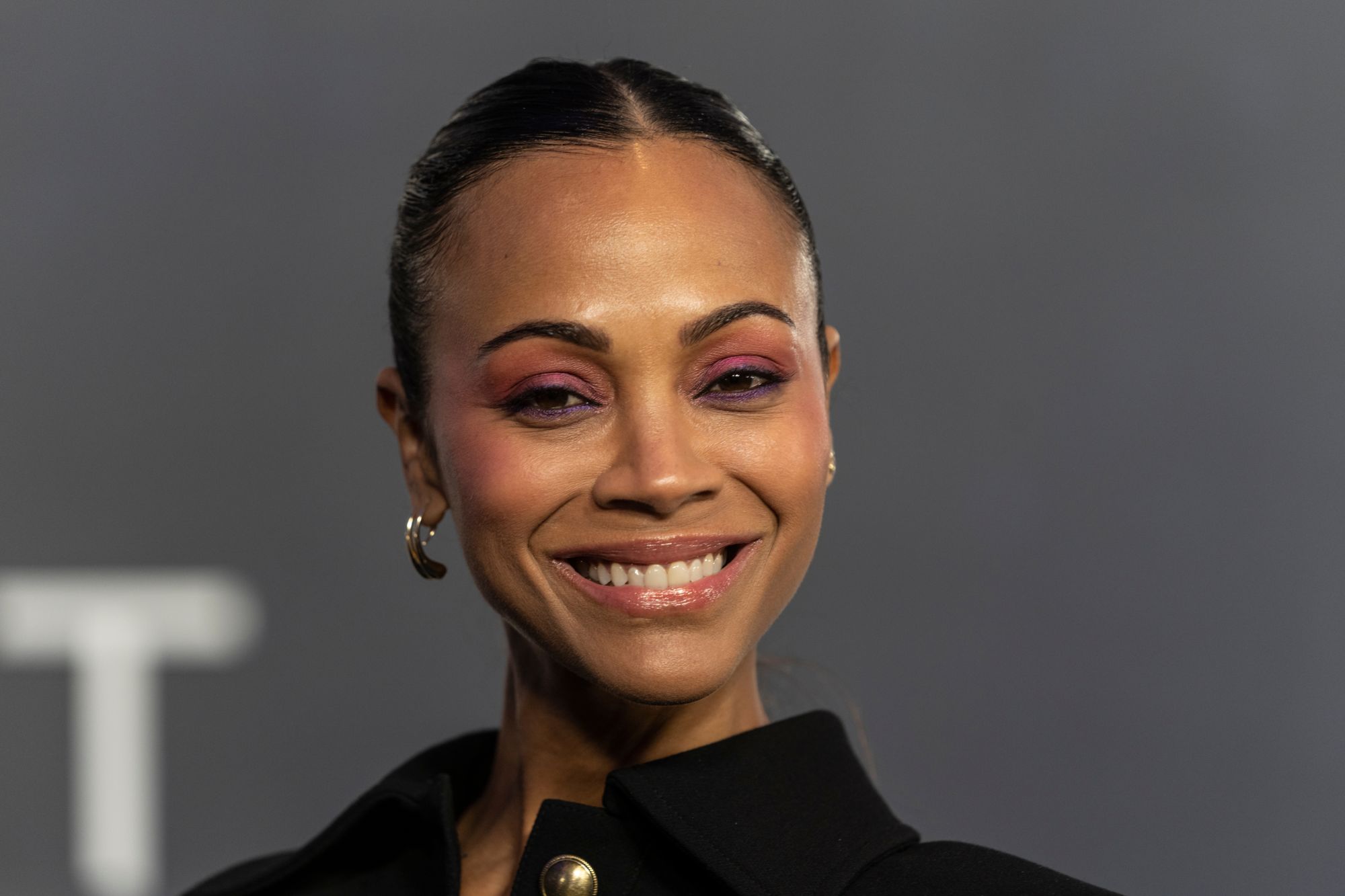Zoe Saldana smiles at a premiere event, showcasing her striking makeup with purples and pinks. She wears a black outfit and hoop earrings, exuding confidence against a gray backdrop.